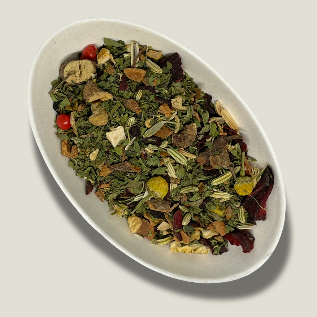 Infusion bio Étreinte d’ambre en vrac dans une coupelle blanche : feuilles de menthe vertes, fleurs de camomille, graines de fenouil, pétales d’hibiscus et baies rouges